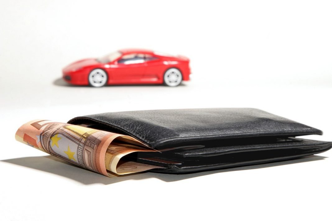 Primer plano de una cartera de la que asoma un fajo de billetes de 50€, con la miniatura de un Ferrari rojo al fondo