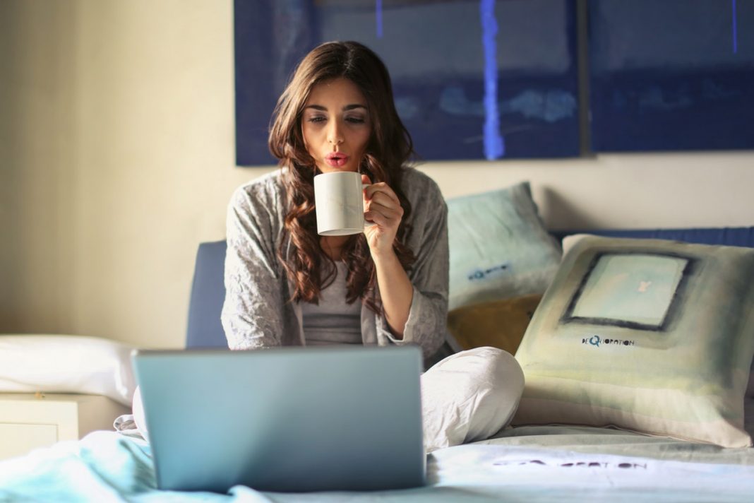 Una mujer está sentada sobre la cama mirando su ordenador y bebiendo de una taza